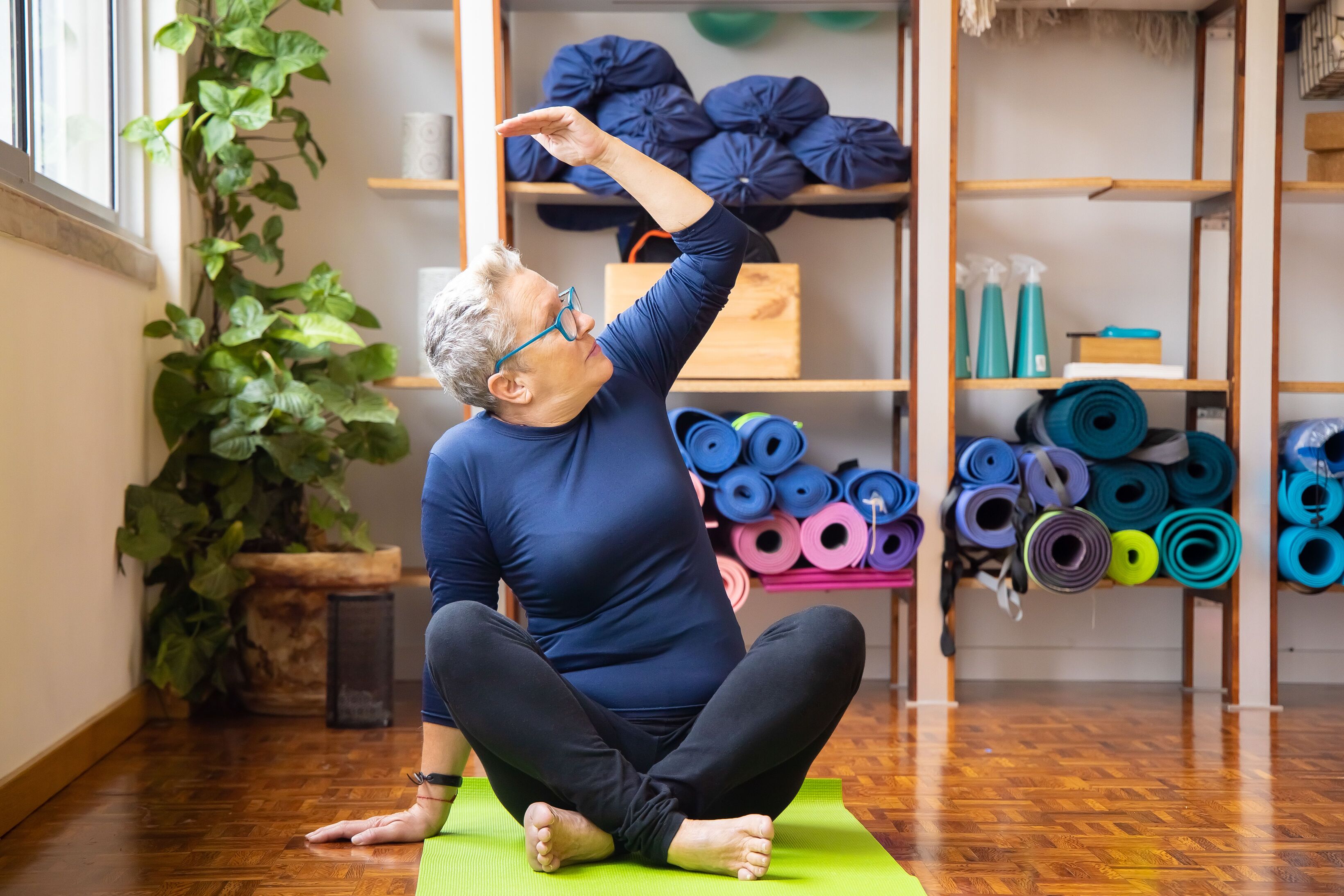 Older woman with grey hair stretching one arm overhead while sitting cross-legged on a mat at home